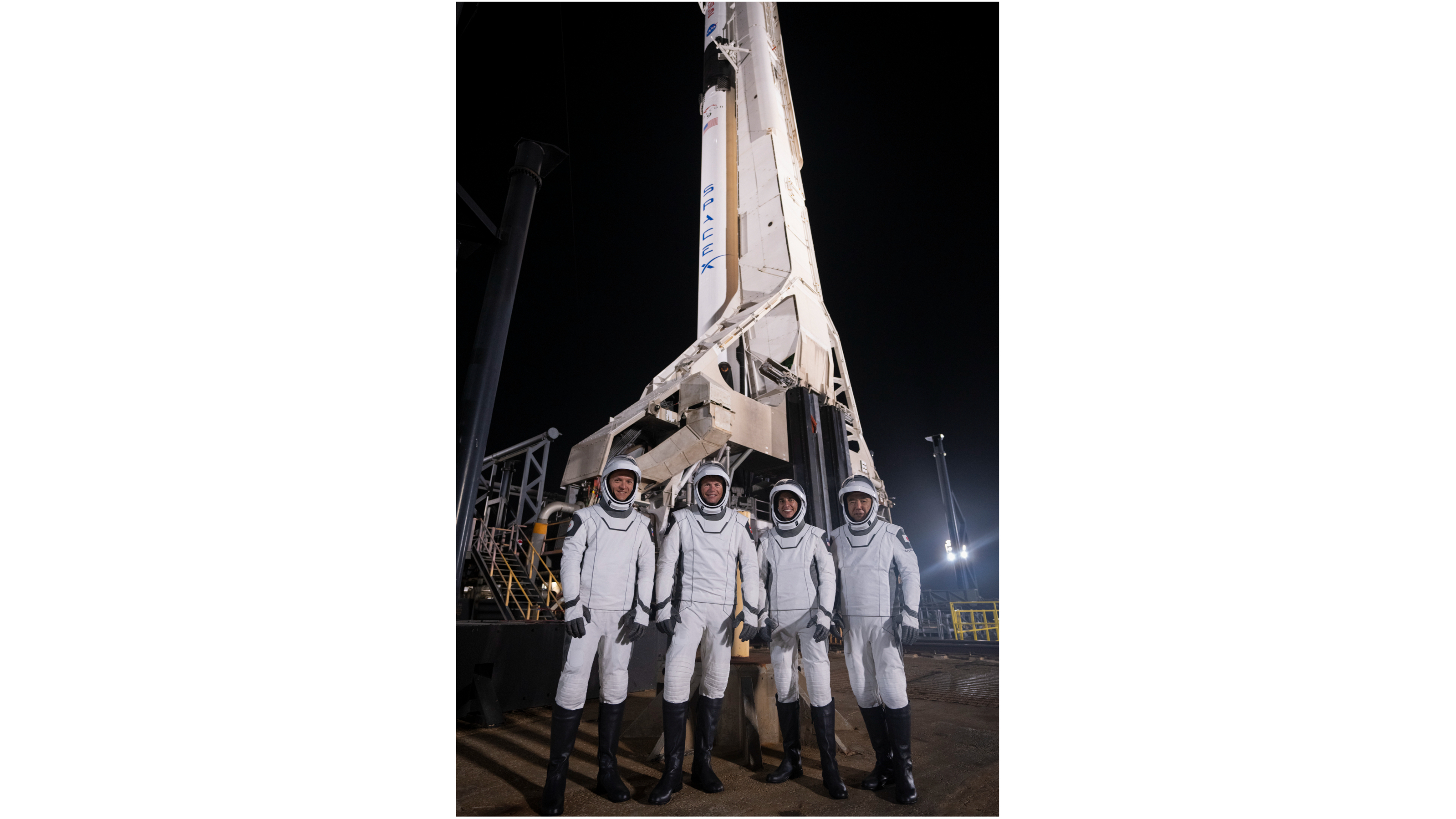 Quatro astronautas em trajes espaciais brancos e pretos ficam em frente a um foguete na plataforma de lançamento à noite.