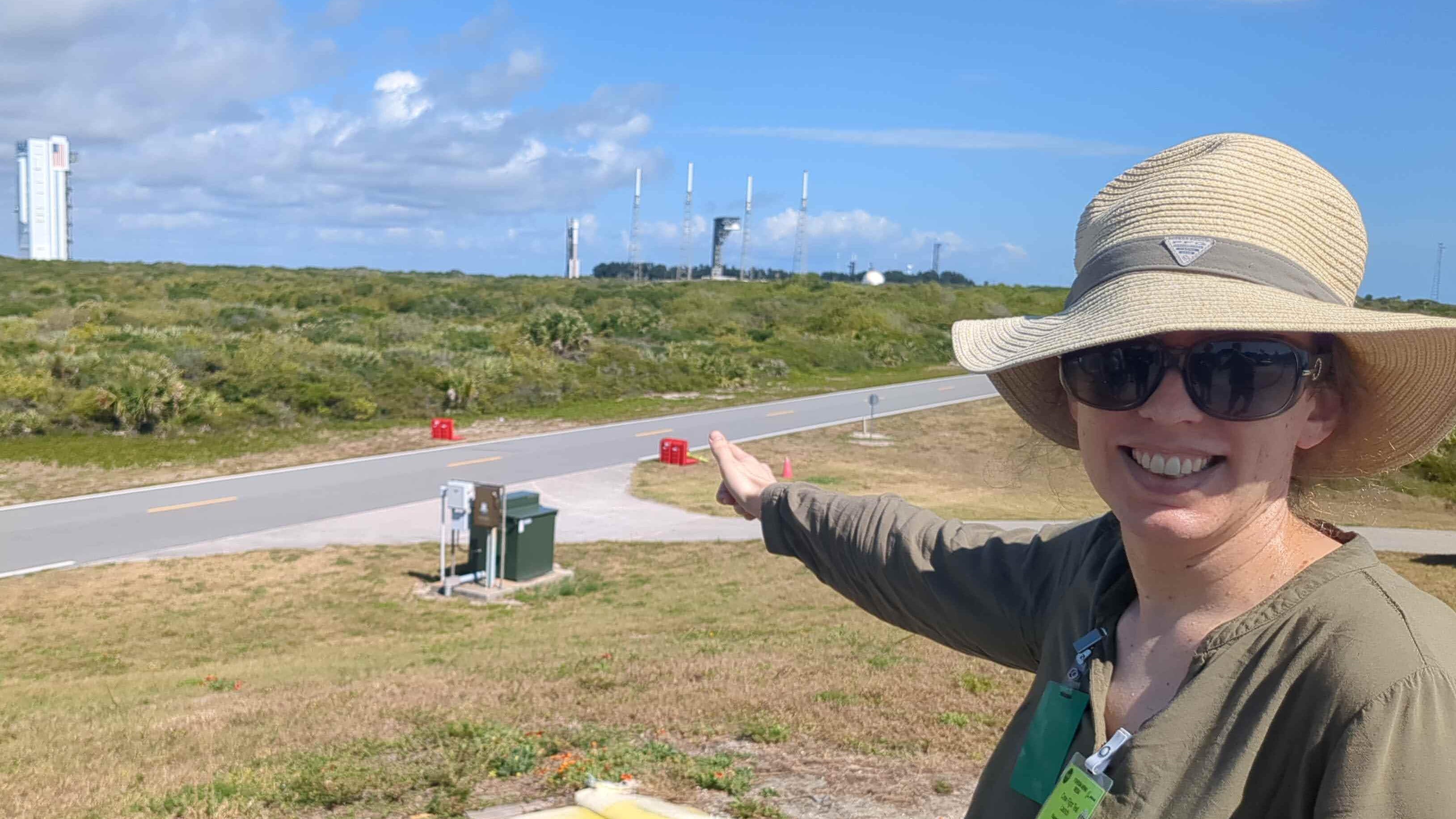 Mulher sorridente, usando um grande chapéu e óculos escuros.  Ela aponta para um foguete do outro lado da estrada e uma plataforma de lançamento distante ao fundo