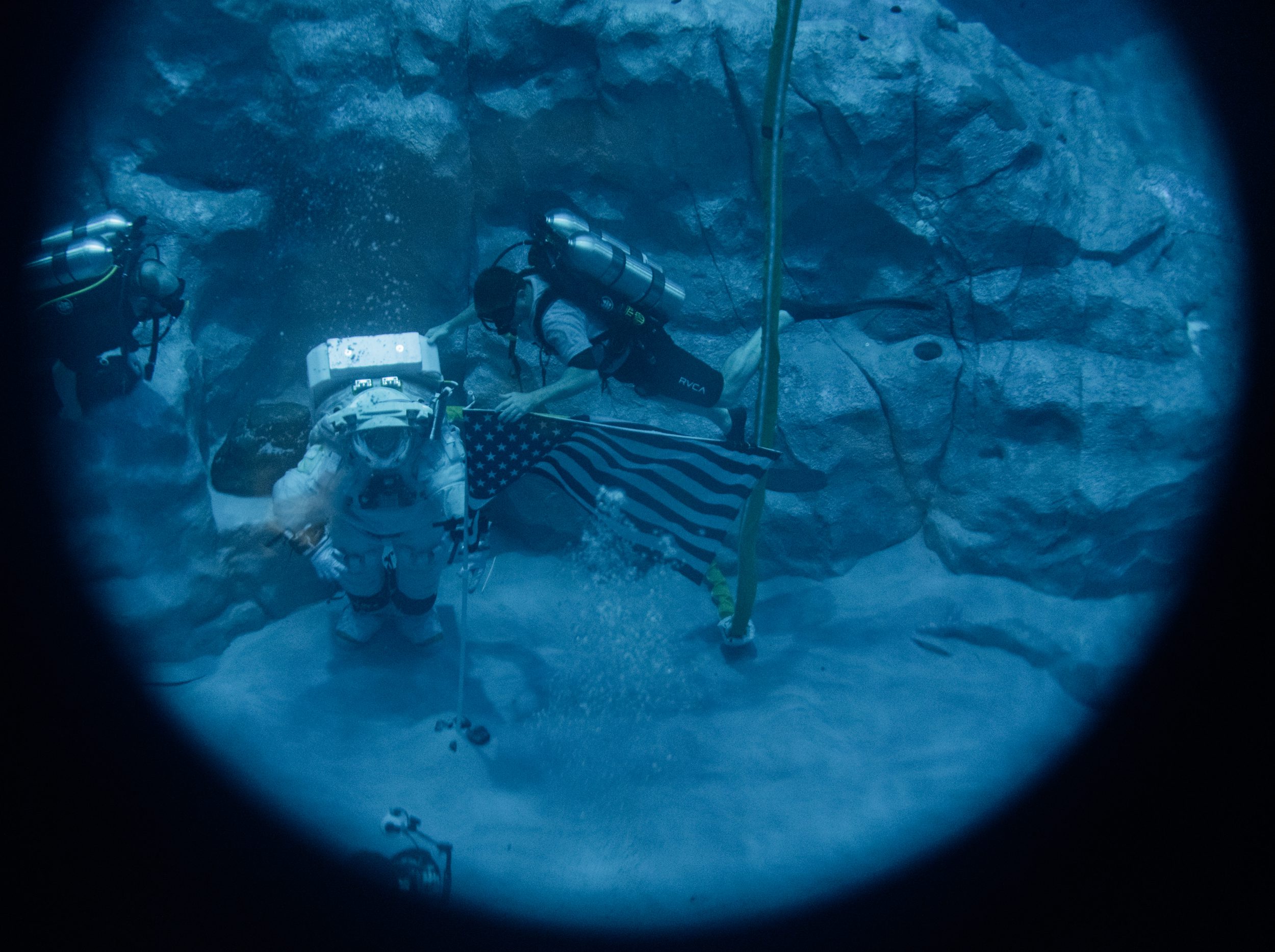 Um astronauta vestindo um traje espacial caminha no fundo de uma piscina ao lado de um mergulhador usando uma máscara de mergulho