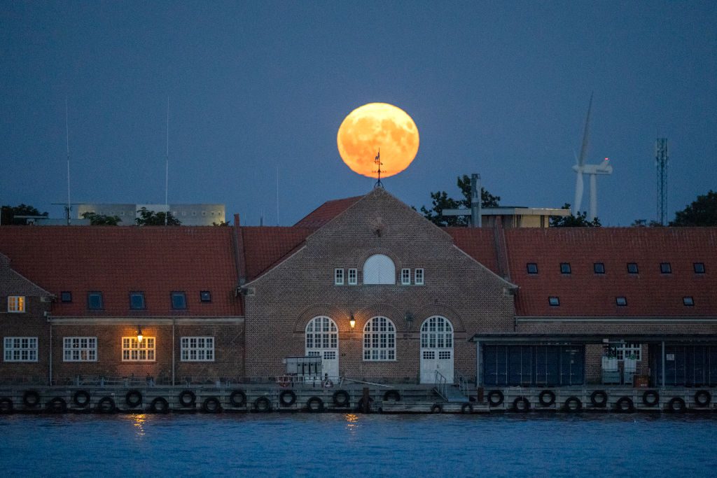 Em primeiro plano está um grande edifício com água na frente e uma grande lua amarelo-alaranjada no céu atrás dele.