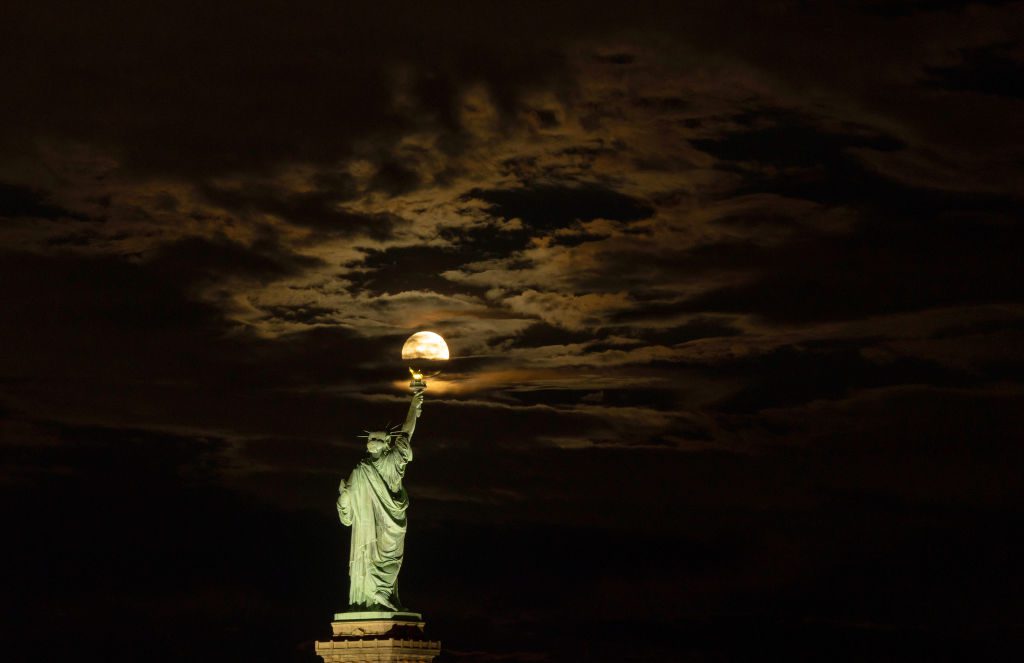 A Estátua da Liberdade está em primeiro plano e a lua brilhante é visível diretamente atrás da estátua através de algumas nuvens.