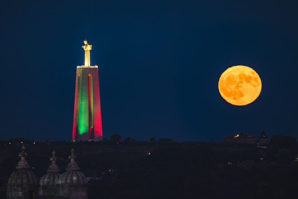 A grande estátua de Cristo à esquerda é iluminada com luzes verdes e vermelhas enquanto a lua cheia brilha intensamente no céu à direita da estátua.